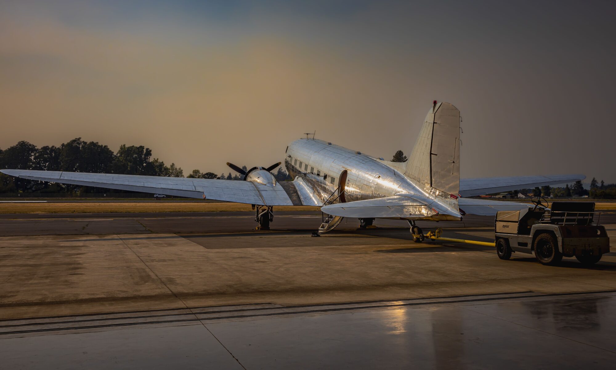 A bare aluminum Douglas DC-3 from the left quartering rear in the evening sunlight, on a tow tug outside a hangar