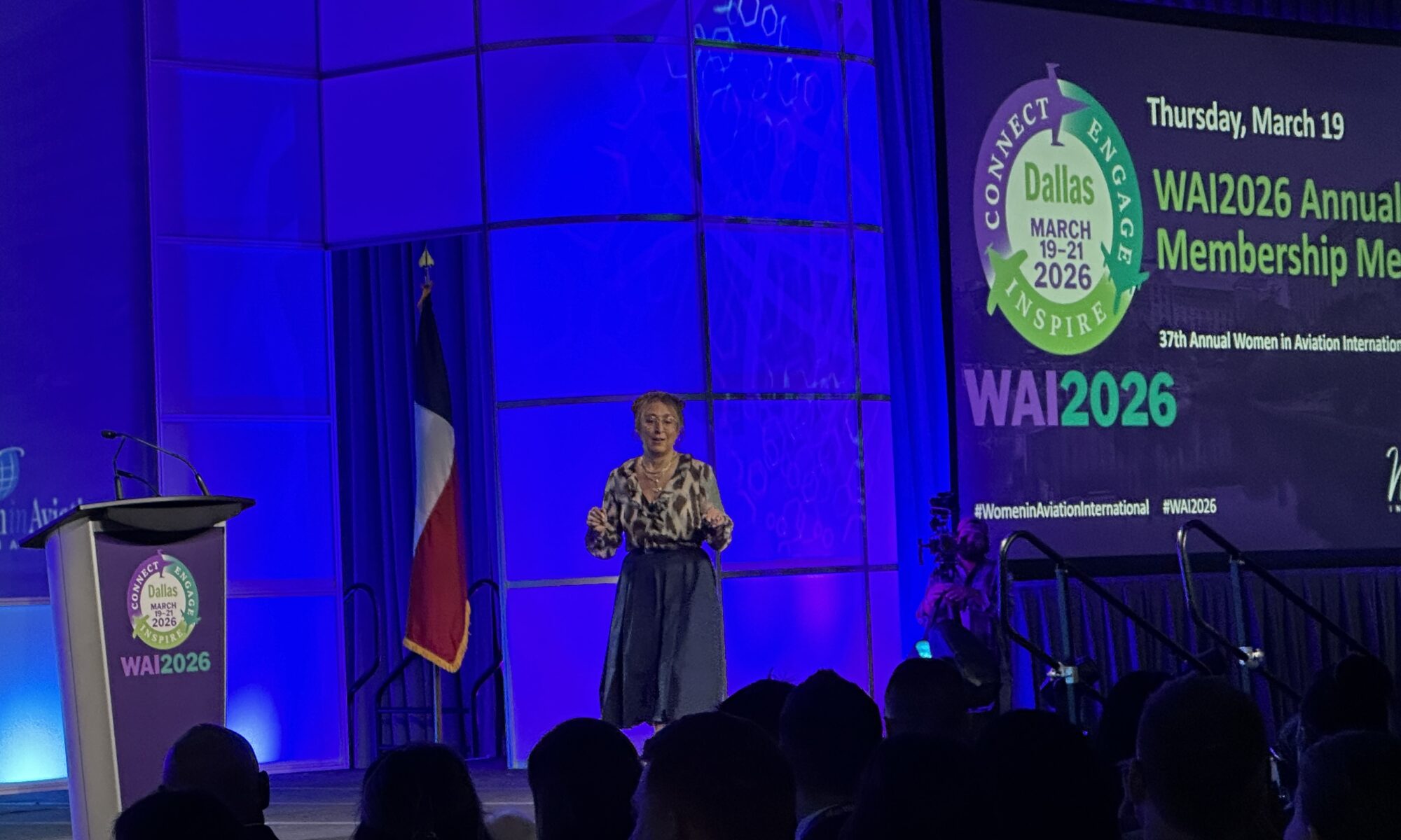 A brunette woman on a stage with deep blue lights in the background and the WAI 26 logo