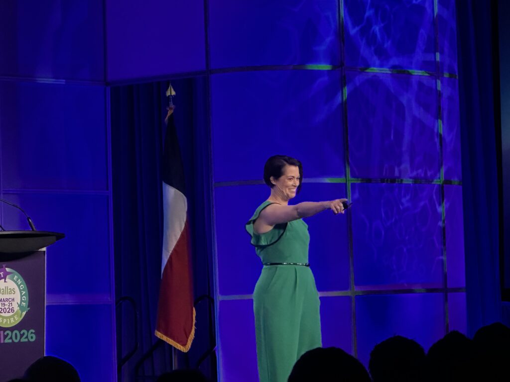 A woman with short dark brunette hair in a kelly green jumpsuit smiles and points to the crowd from the stage