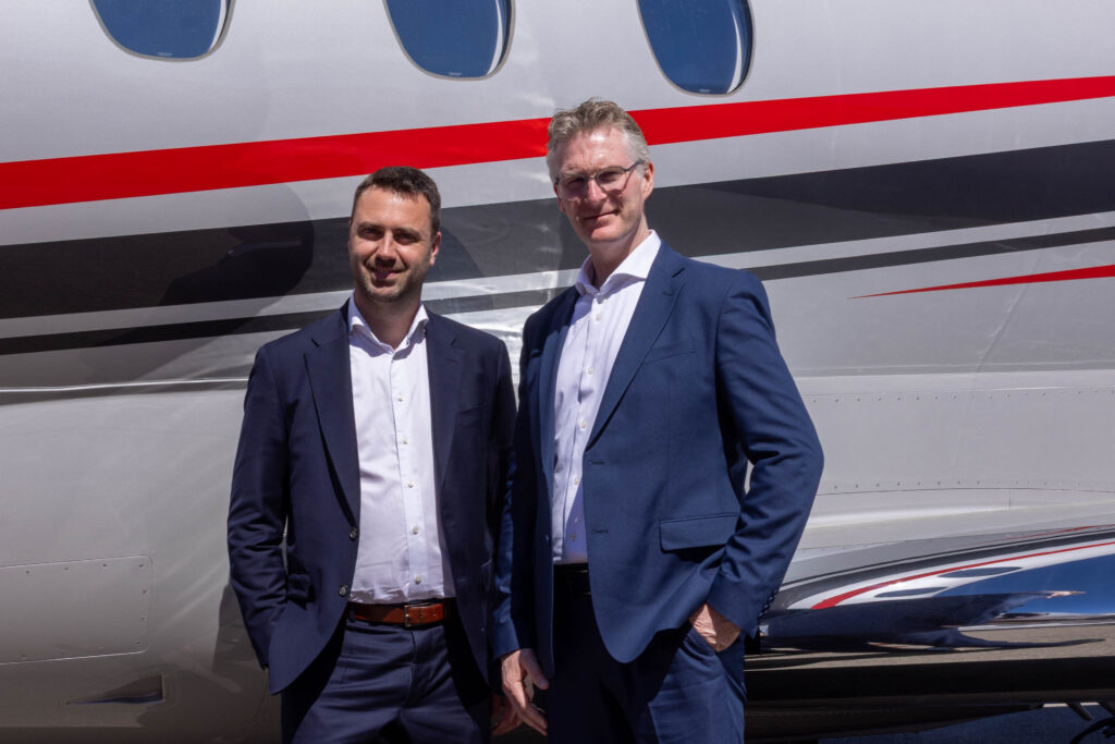 two men in dark blue suits in front of a gray jet with red and black stripes