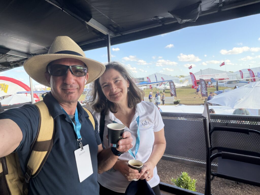 a man in a straw hat and a woman in a white polo on an RV porch with cups of coffee