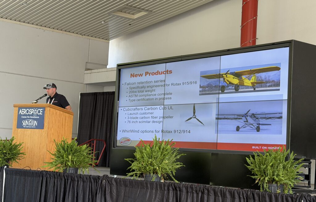 a man behind a pale wooden lecturn next to a large screen with New Products and airplanes on it