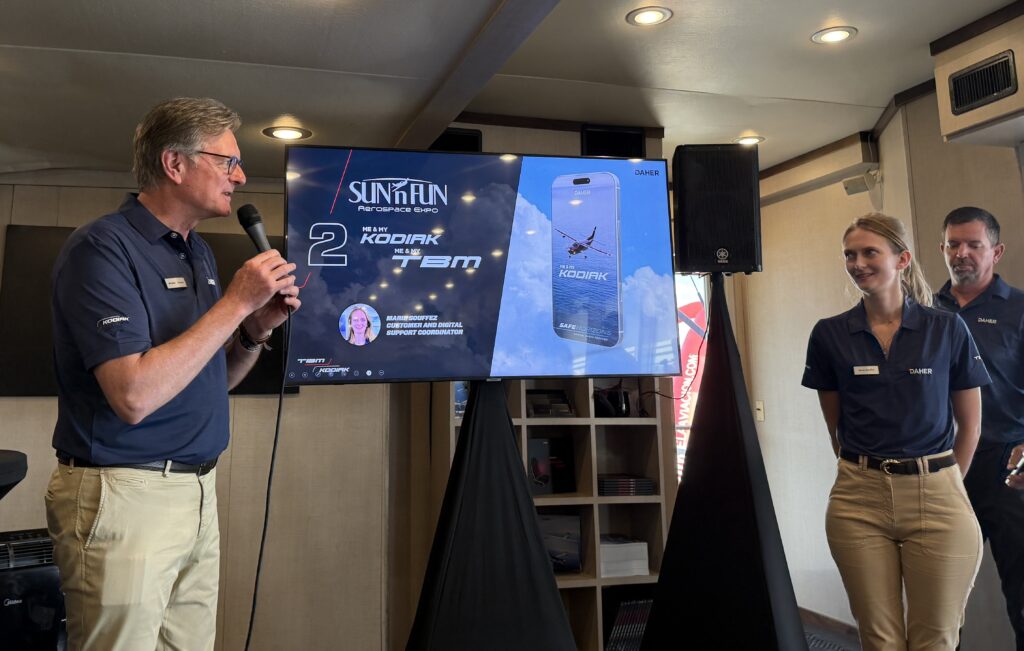 a tall man in profile with a microphone introduces a young woman with a ponytail in front of a screen