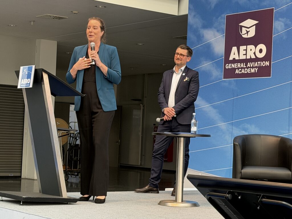 A woman at a lecturn speaks into a mike with a man with a goatee and glasses behind her.