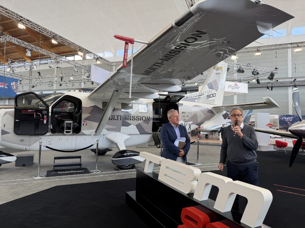 Two men stand in front of a high wing utility Kodiak 900 in gray camo paint, with a TBM 980 podium in front.