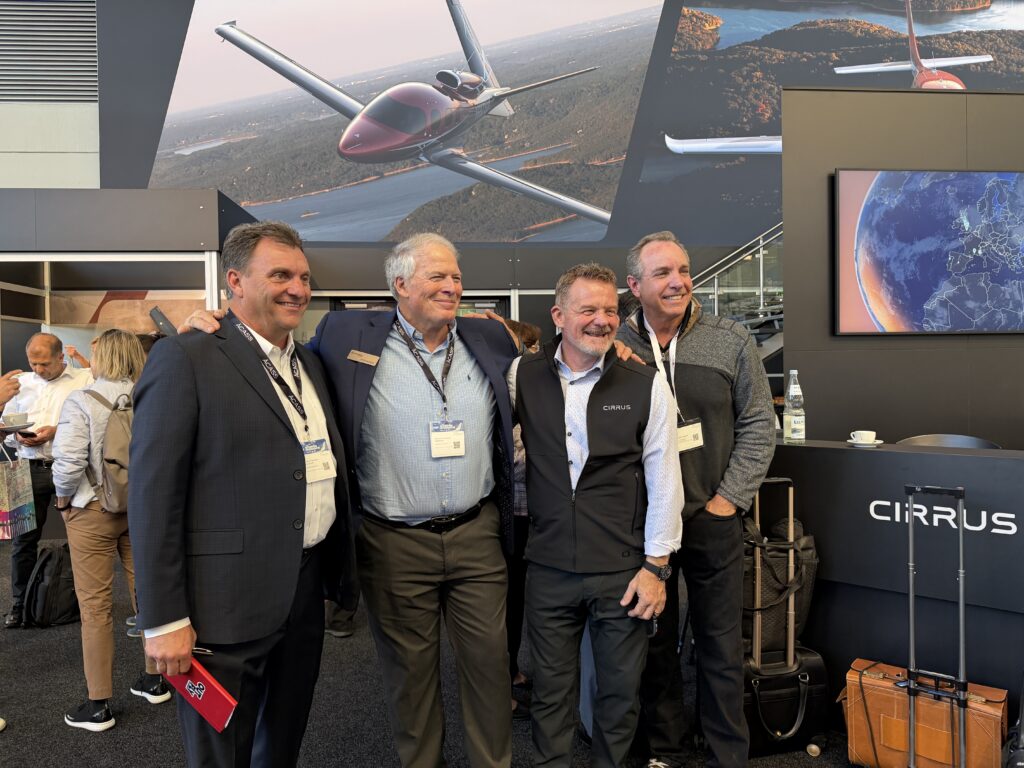 Four middle aged white men stand in front of a banner with a Cirrus Vision Jet on it.