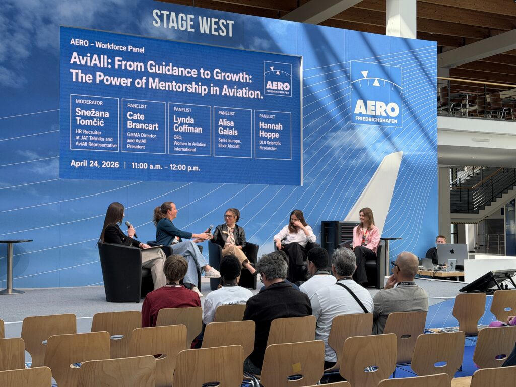 A group of five women sit in comfy chairs on a stage in front of a blue banner
